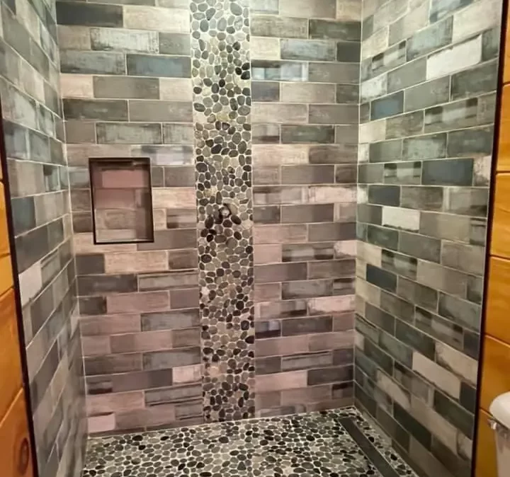 A walk in shower with light blue-green and some off white small format tiles installed on a 50% pattern on the walls with same colored pebble sized stones on the floor and as a 12 inch wide accent going vertical in the middle of the back wall. There is a 12 by 24 inch niche installed vertically on the left side of the accent near chest height